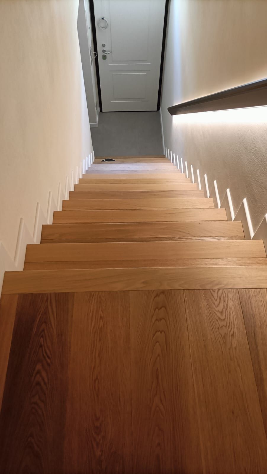 Scala in legno che scende verso una porta chiusa, con un corrimano illuminato lungo la parete destra.