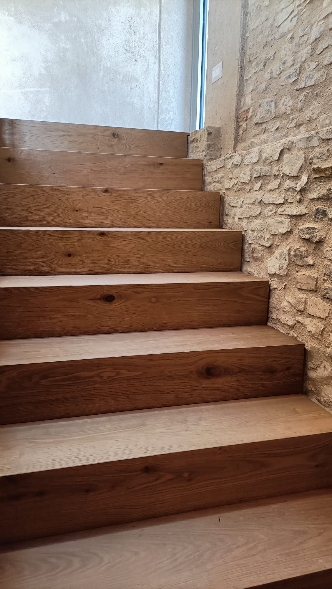 Scala in legno con alzate bianche accanto a una parete in pietra lavorata, illuminata dalla luce naturale proveniente dall'alto.