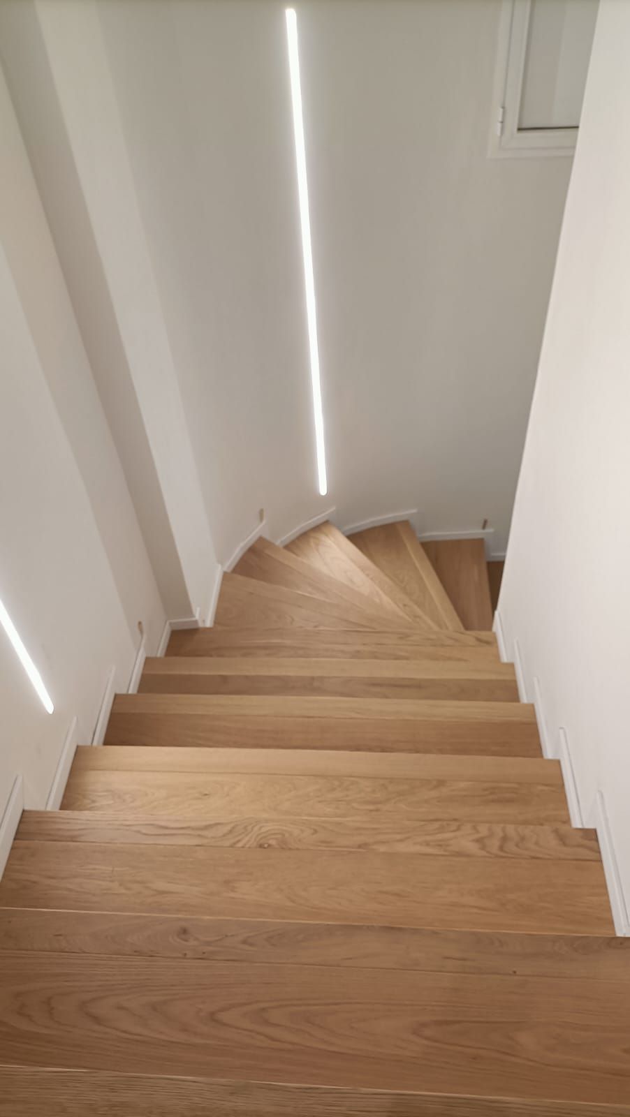 Una scala di legno scende in uno stretto corridoio bianco illuminato da una fessura luminosa sulla parete.