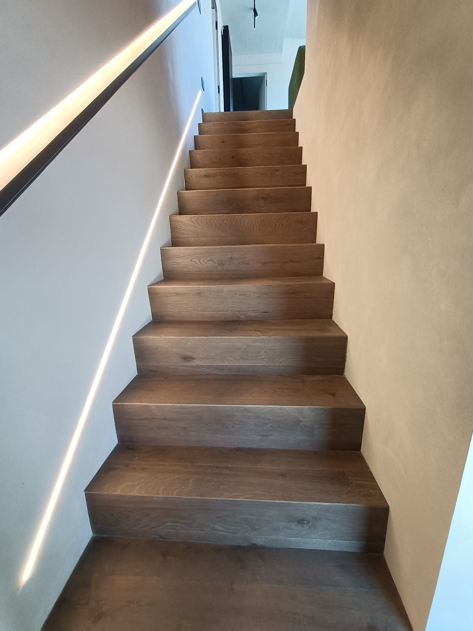 Scala interna ripida con gradini in legno, parete bianca e corrimano sulla sinistra.