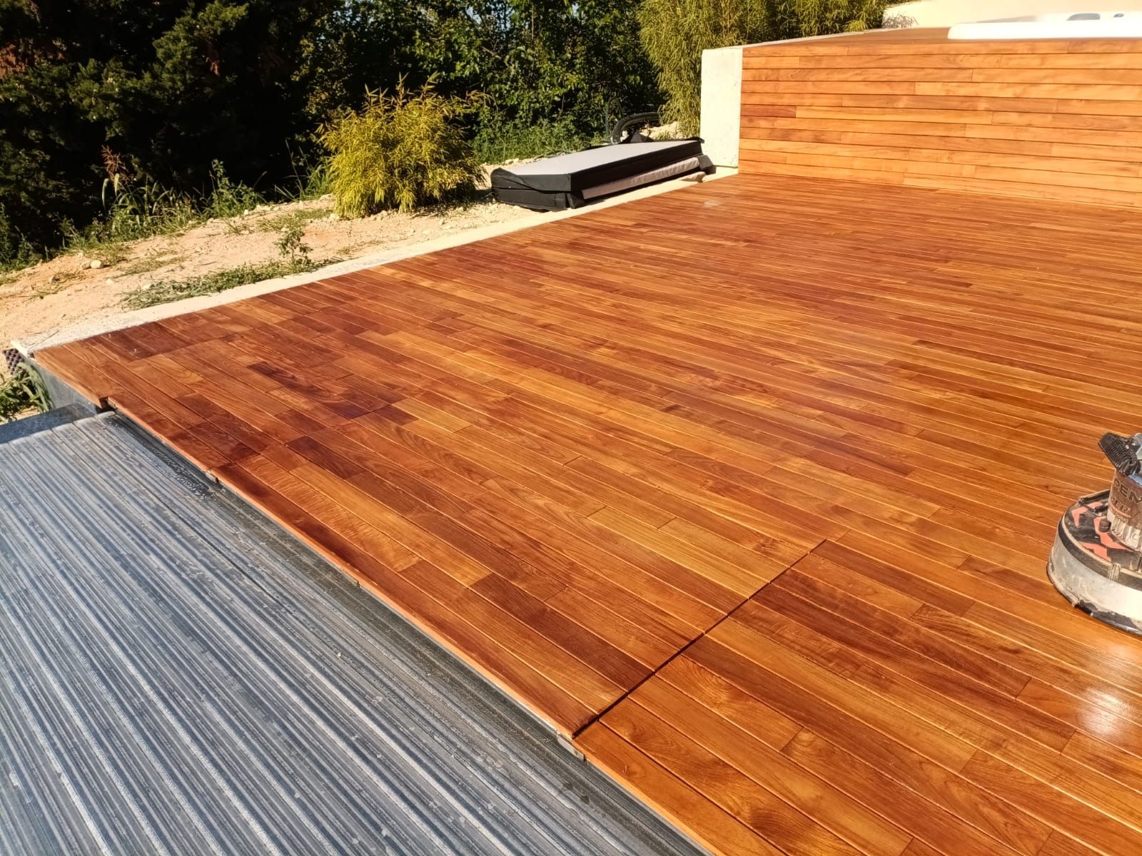 Terrazza in legno che viene tinta con una levigatrice, accanto a una terrazza grigia non rifinita e a un muro di mattoni.