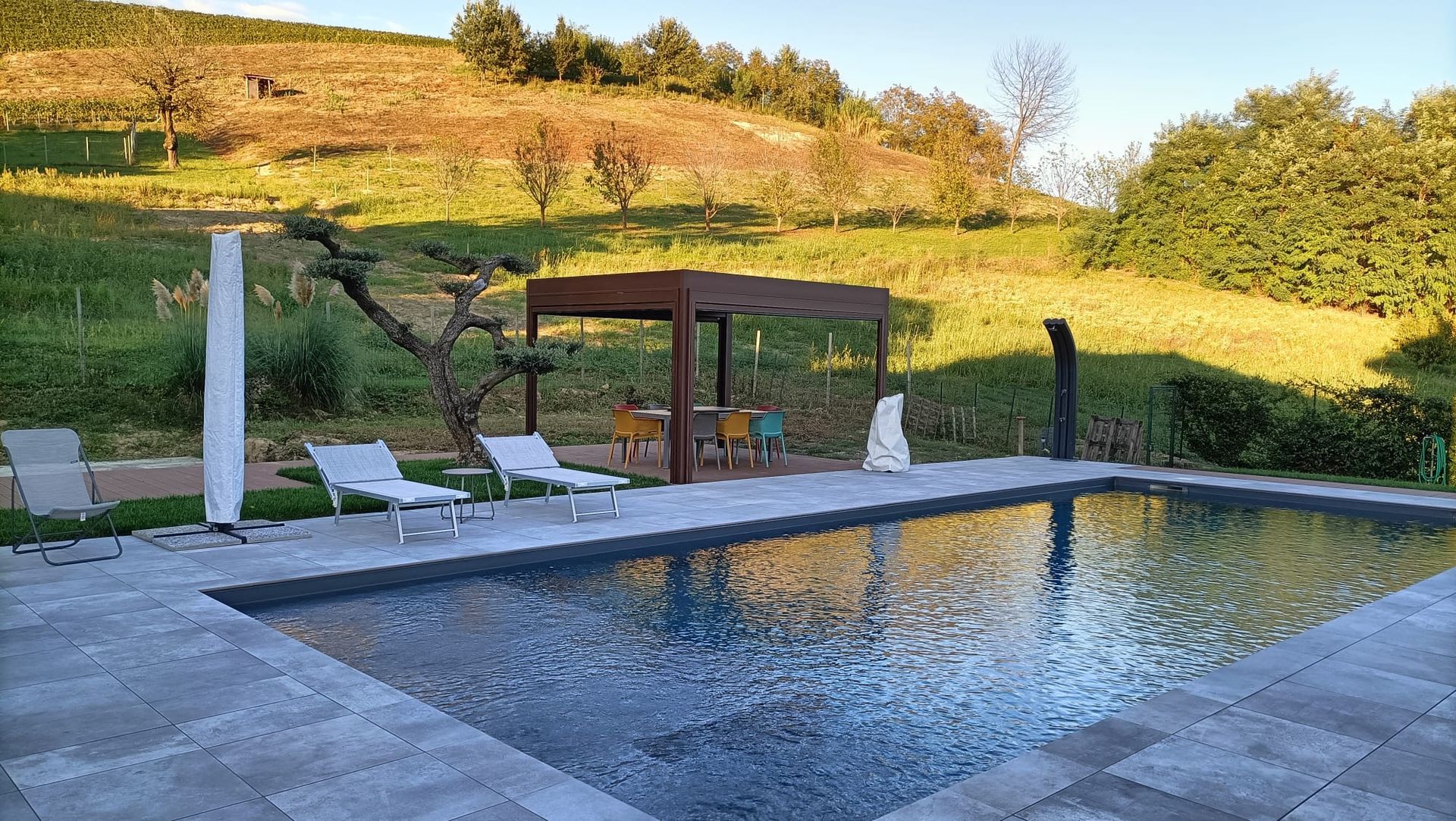 Piscina moderna in giardino, accanto a lettini prendisole e un gazebo ombreggiato, con una collina erbosa sullo sfondo.