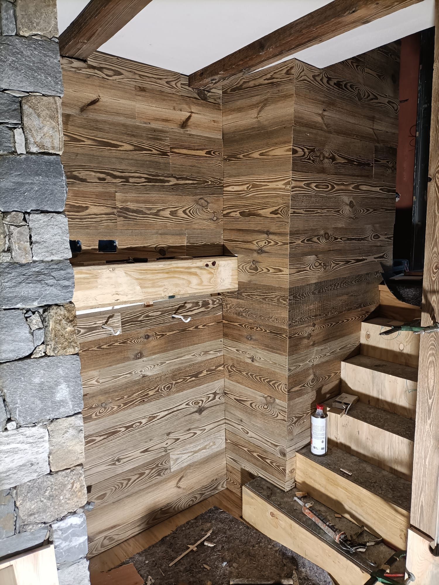 Angolo interno rivestito in legno con travi a vista, muro in pietra grezza e gradini non rifiniti.