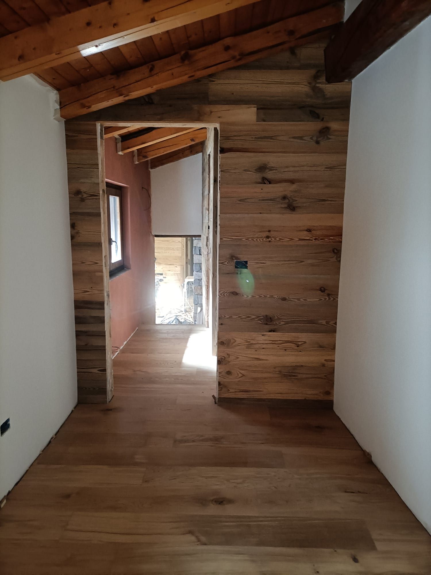 Corridoio rustico con soffitto in legno, pareti bianche e una porta che conduce a una stanza luminosa