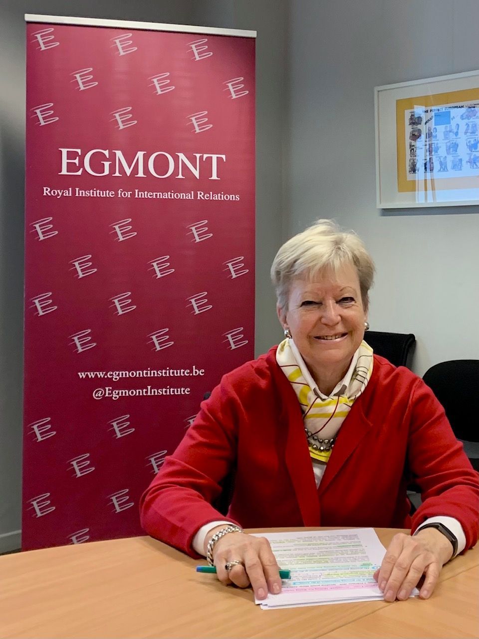 Eine lächelnde Person in einem roten Blazer sitzt an einem Schreibtisch mit Papieren vor einem Banner des Egmont-Instituts.