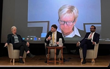 Ein dreiköpfiges Panel sitzt auf der Bühne, hinter ihnen wird auf einer großen Leinwand eine vierte Person gezeigt.
