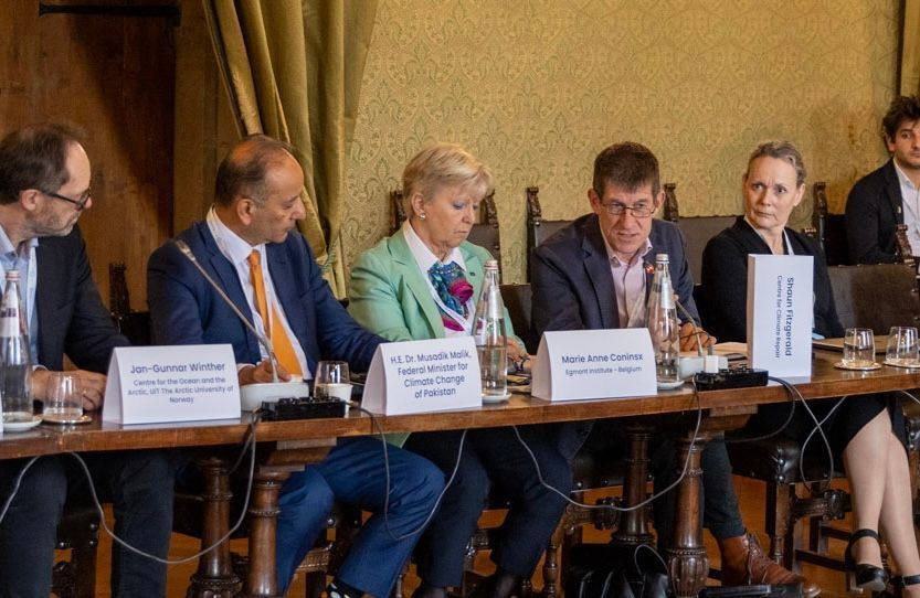 Fünf Personen sitzen während einer formellen Besprechung an einem langen Holztisch, jede mit einem Namensschild vor sich.