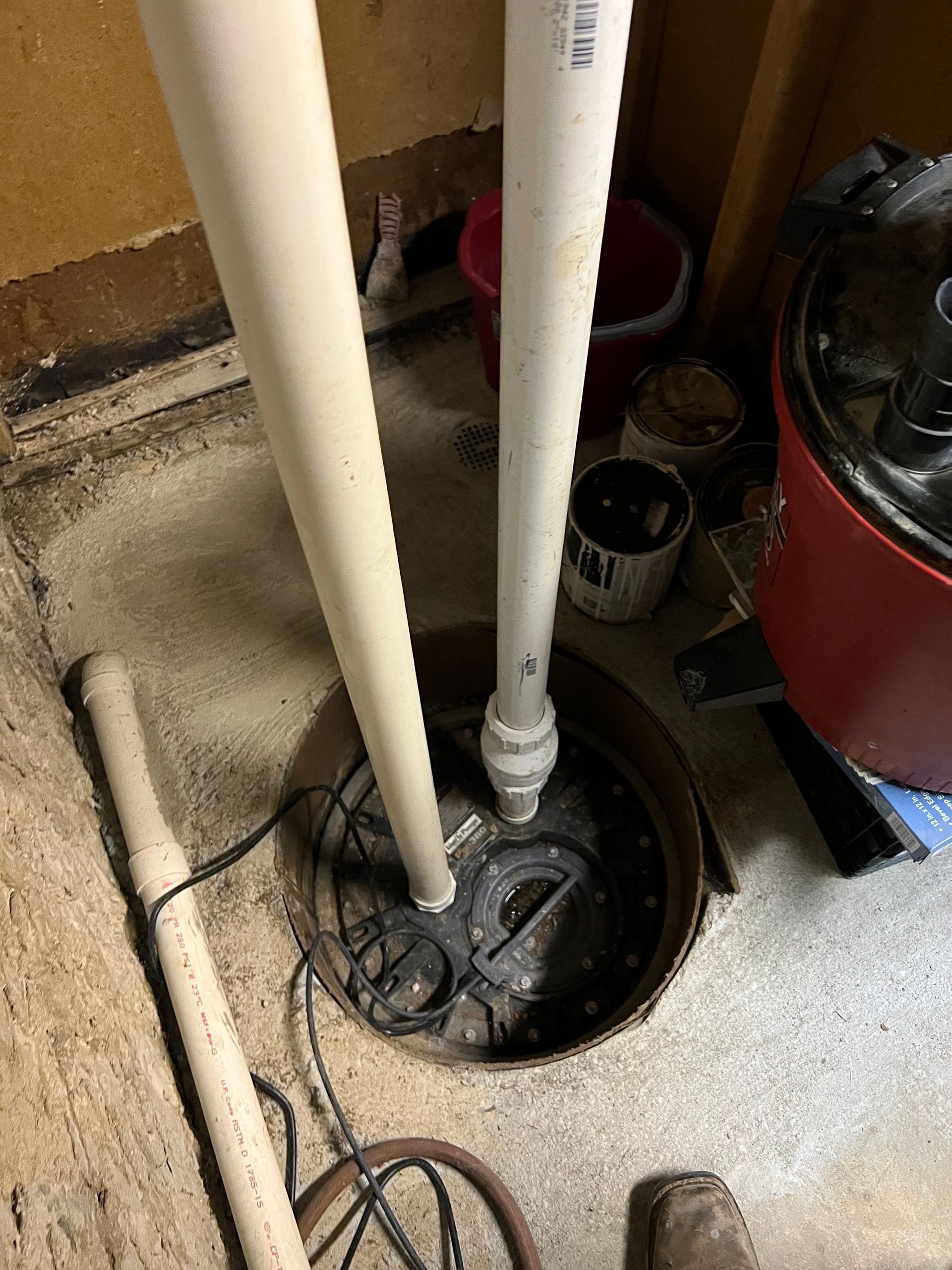 Sump pump pit with two white pipes, electrical cords, and a red appliance in a basement.