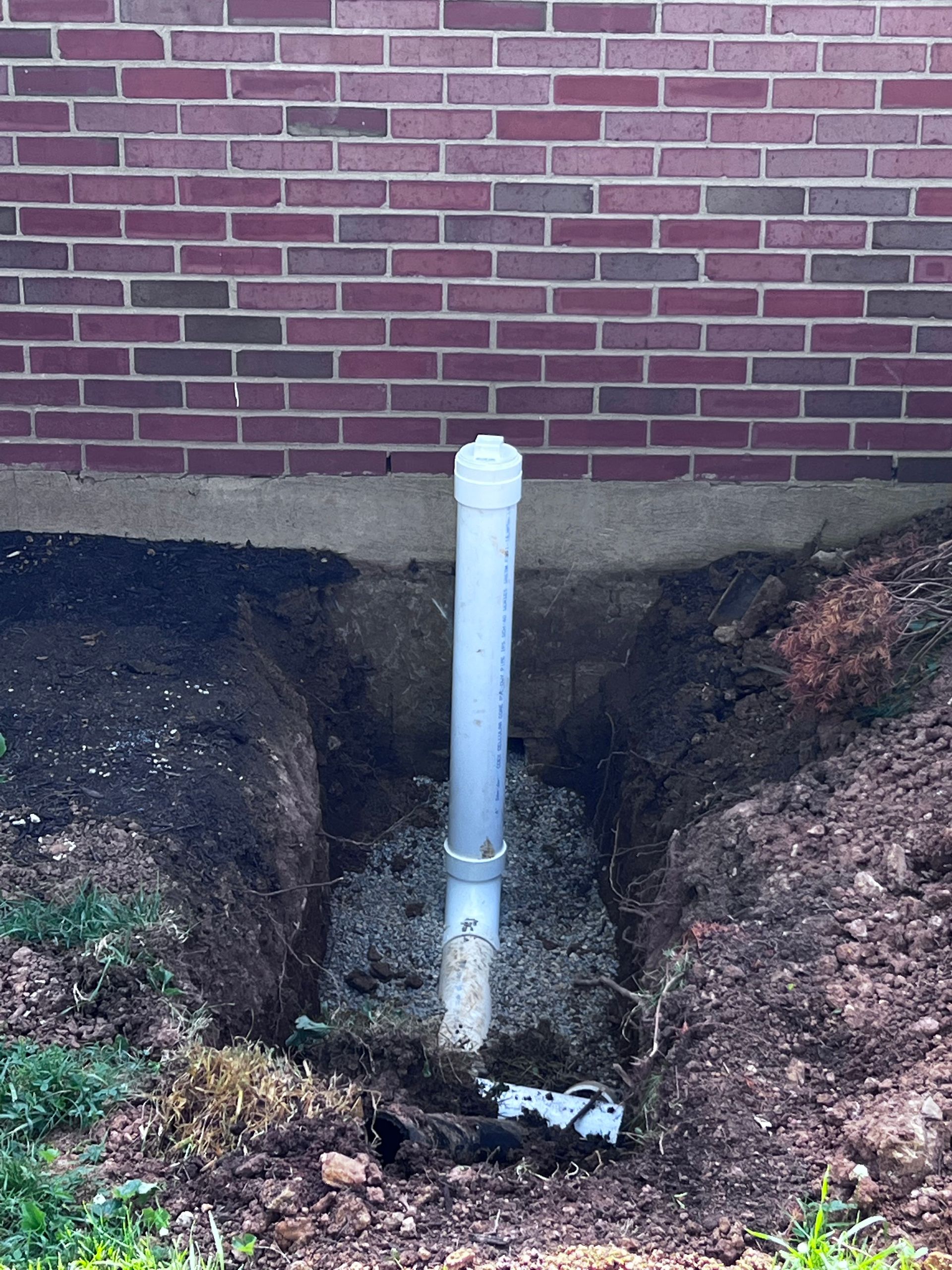 A PVC pipe in a trench beside a brick building. The trench is filled with gravel and dirt.