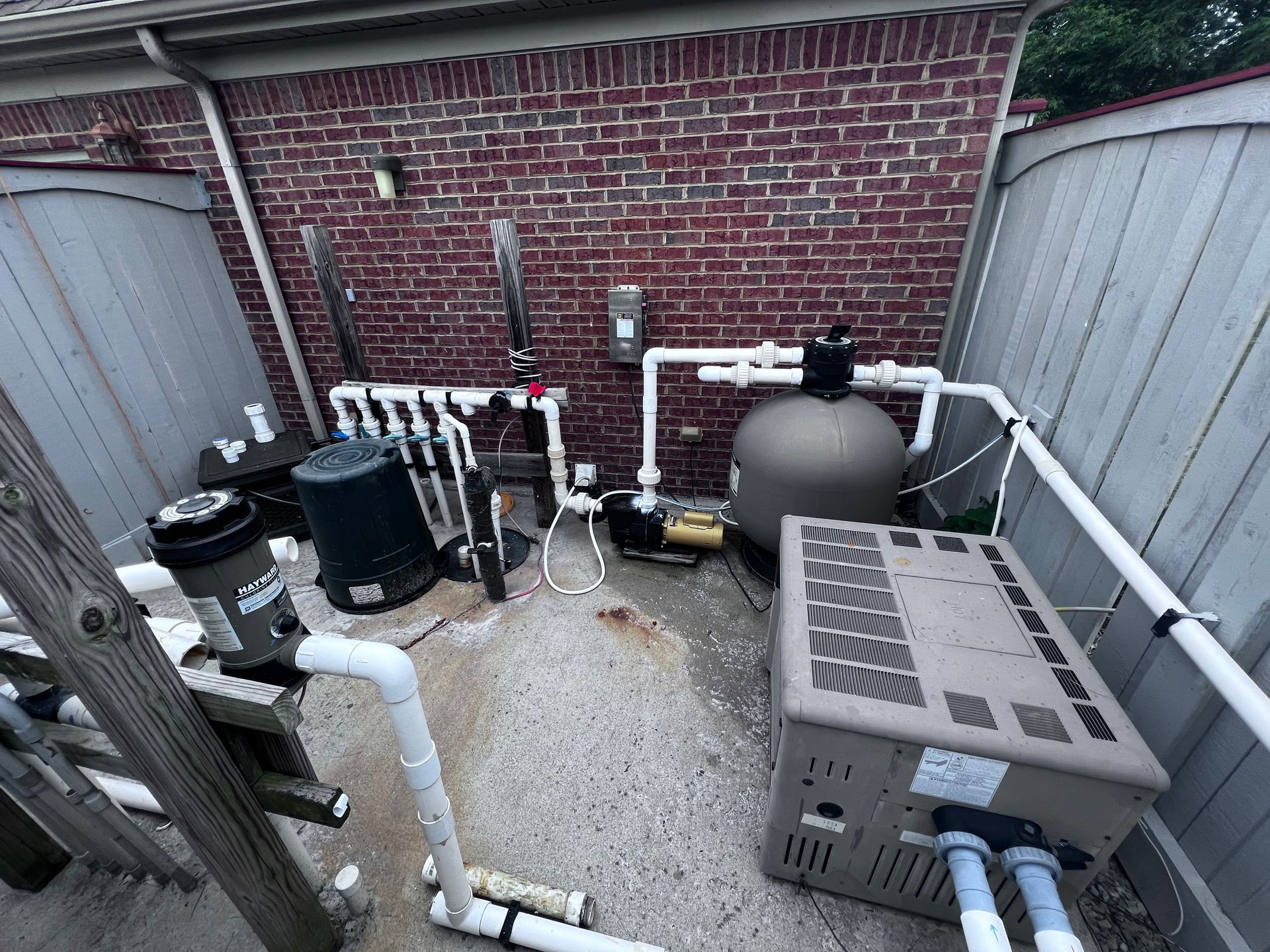 Pool equipment, including a filter, pump, and heater, set against a brick wall and wooden fence.