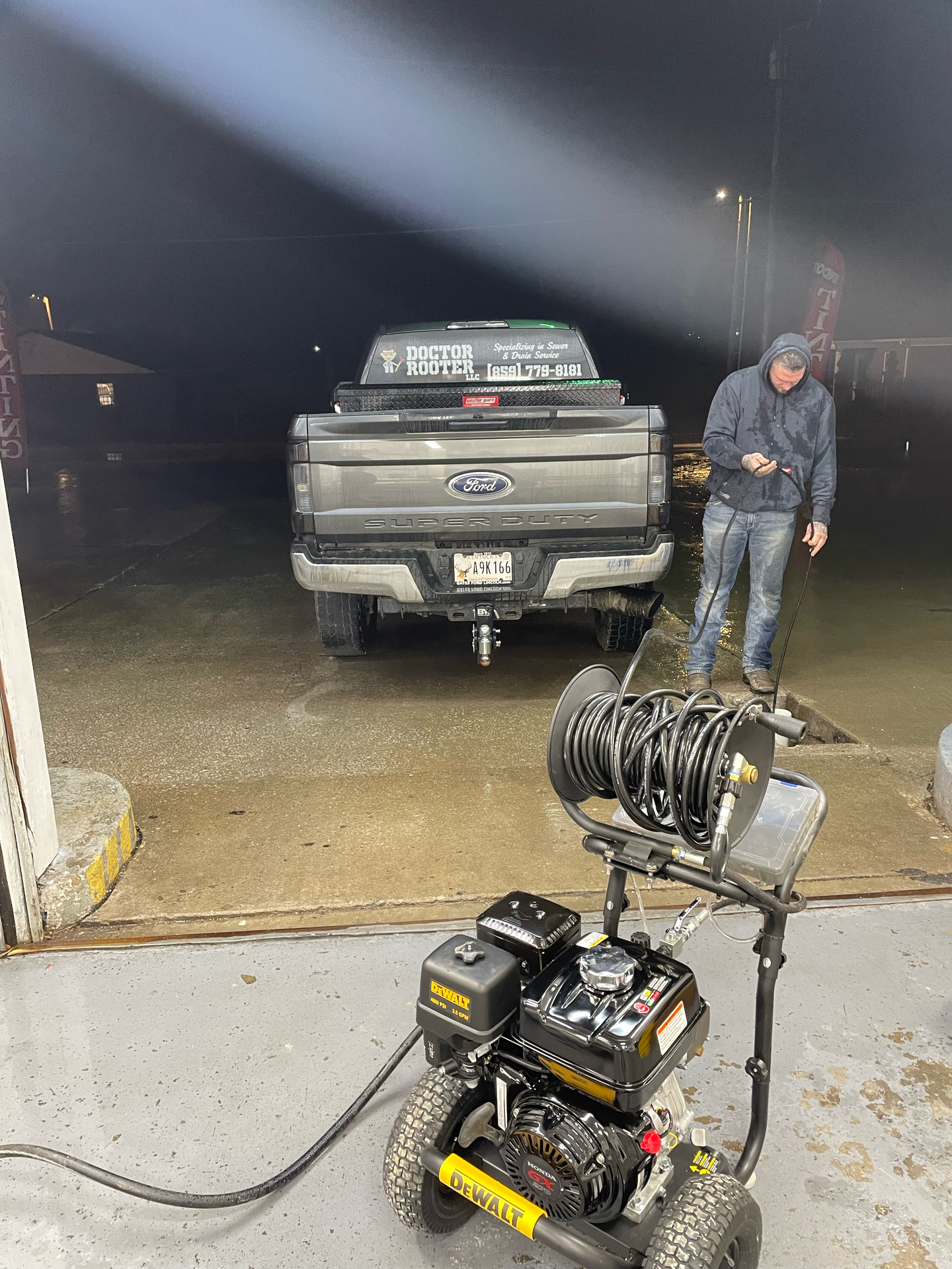 A man uses a pressure washer on a gray pickup truck at a car wash. It's night, wet.