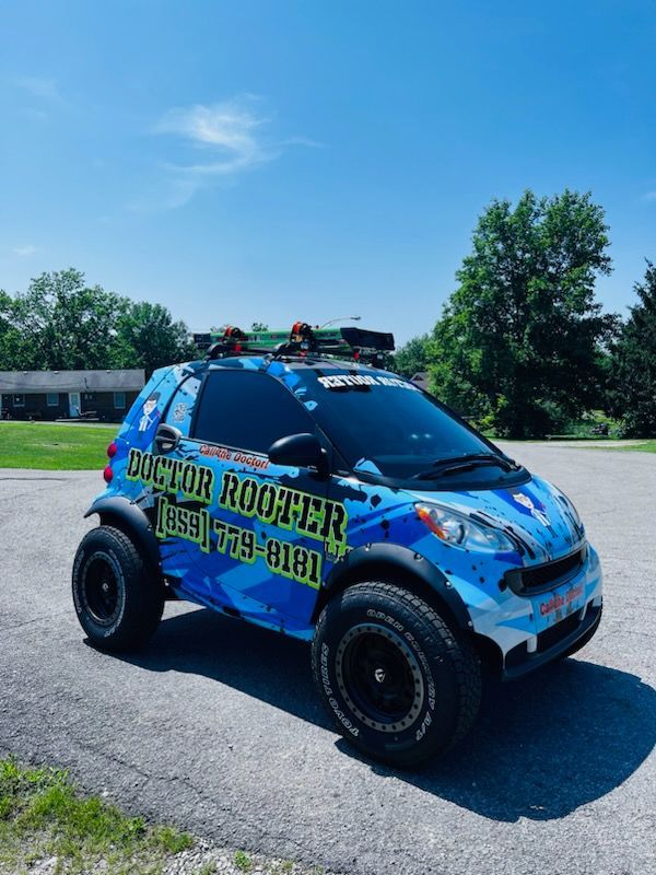 Modified Smart car with blue wrap, large tires, and Doctor Roofer logo parked on pavement under a blue sky.