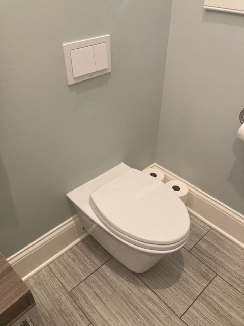 White wall-mounted toilet in a small bathroom with gray tiled floor and flush buttons above.