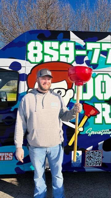 Man in a gray hoodie holding a large plunger stands in front of a blue van with plumbing company logo and phone number.