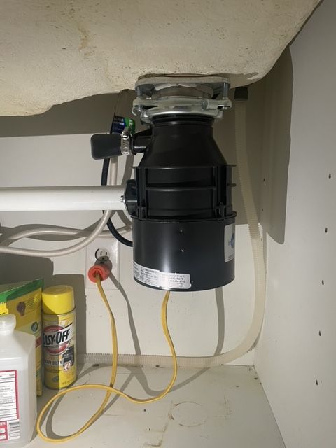 Garbage disposal unit mounted under a kitchen sink. Black and silver appliance with yellow power cord.