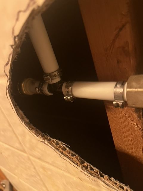 Pipes connected by fittings, visible through a hole in a wall. White and black pipes with metal clamps are shown.