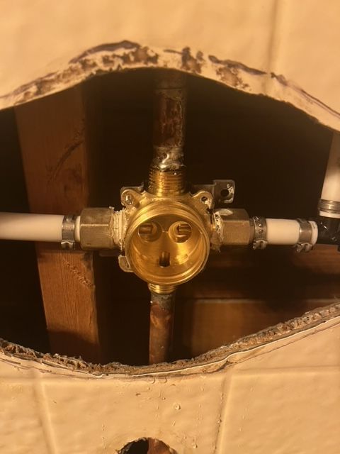 Shower valve fixture in a wall cutout. Brass-colored valve with three white pipes and one copper pipe attached.