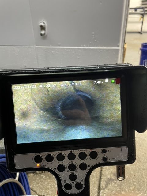 Camera screen showing a close-up view inside a pipe. Dark, circular hole with brown and blue coloration.