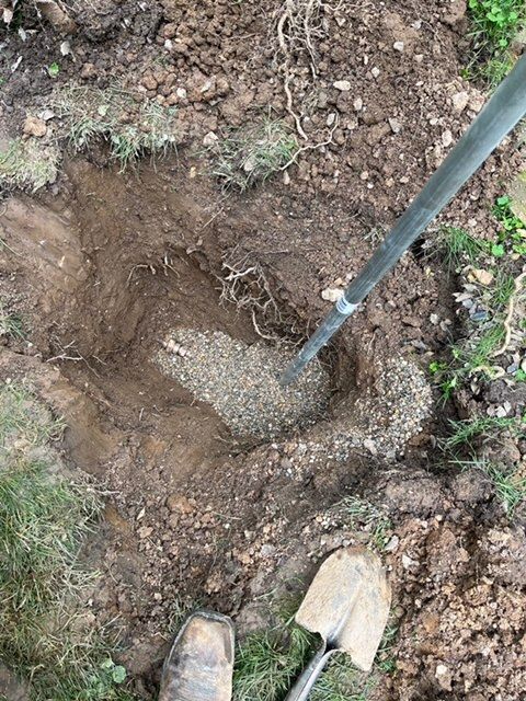 Person digging a hole in the ground, gravel visible at the bottom. Shovel and boot in view.