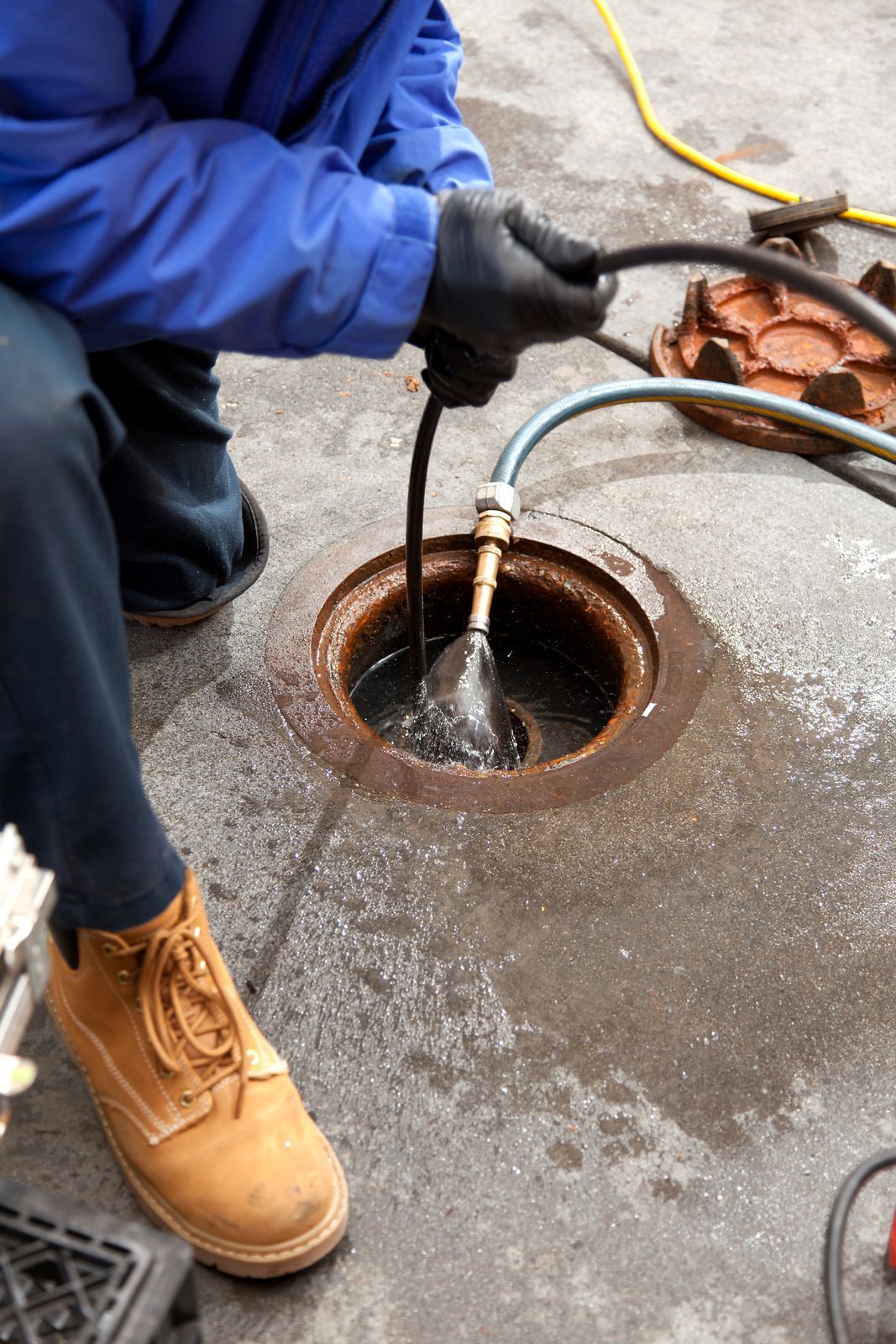 Person in blue overalls and gloves cleaning a drain with a hose.