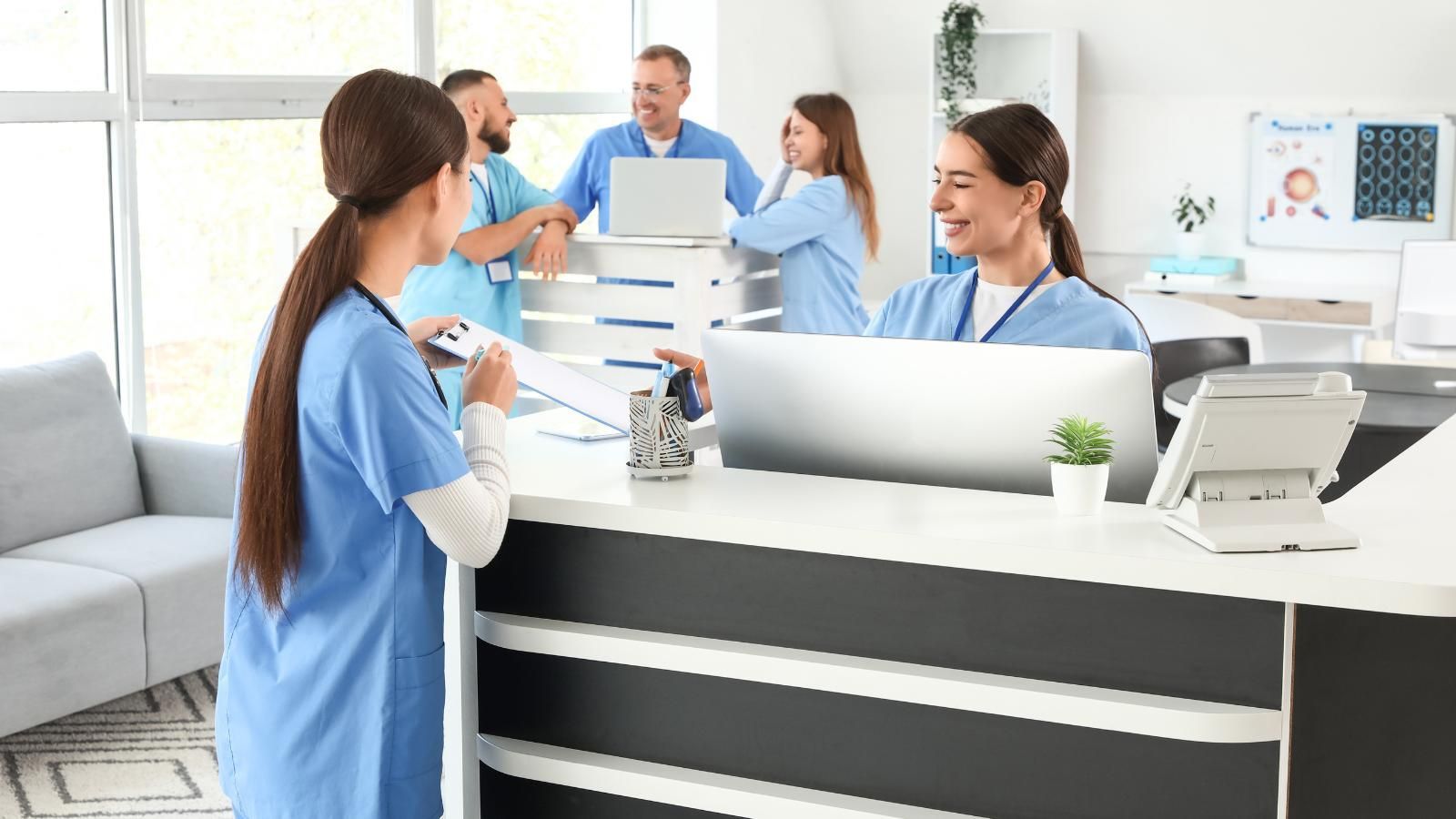 Medical staff at a reception desk, one taking notes, others behind the counter talking near laptops, bright office.