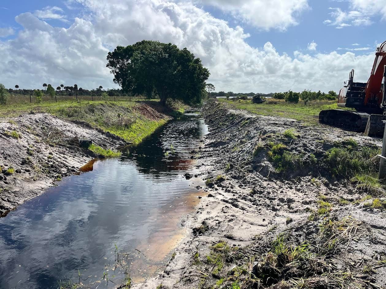 Water-filled drainage ditch in a rural field under a cloudy sky, with an excavator at right.