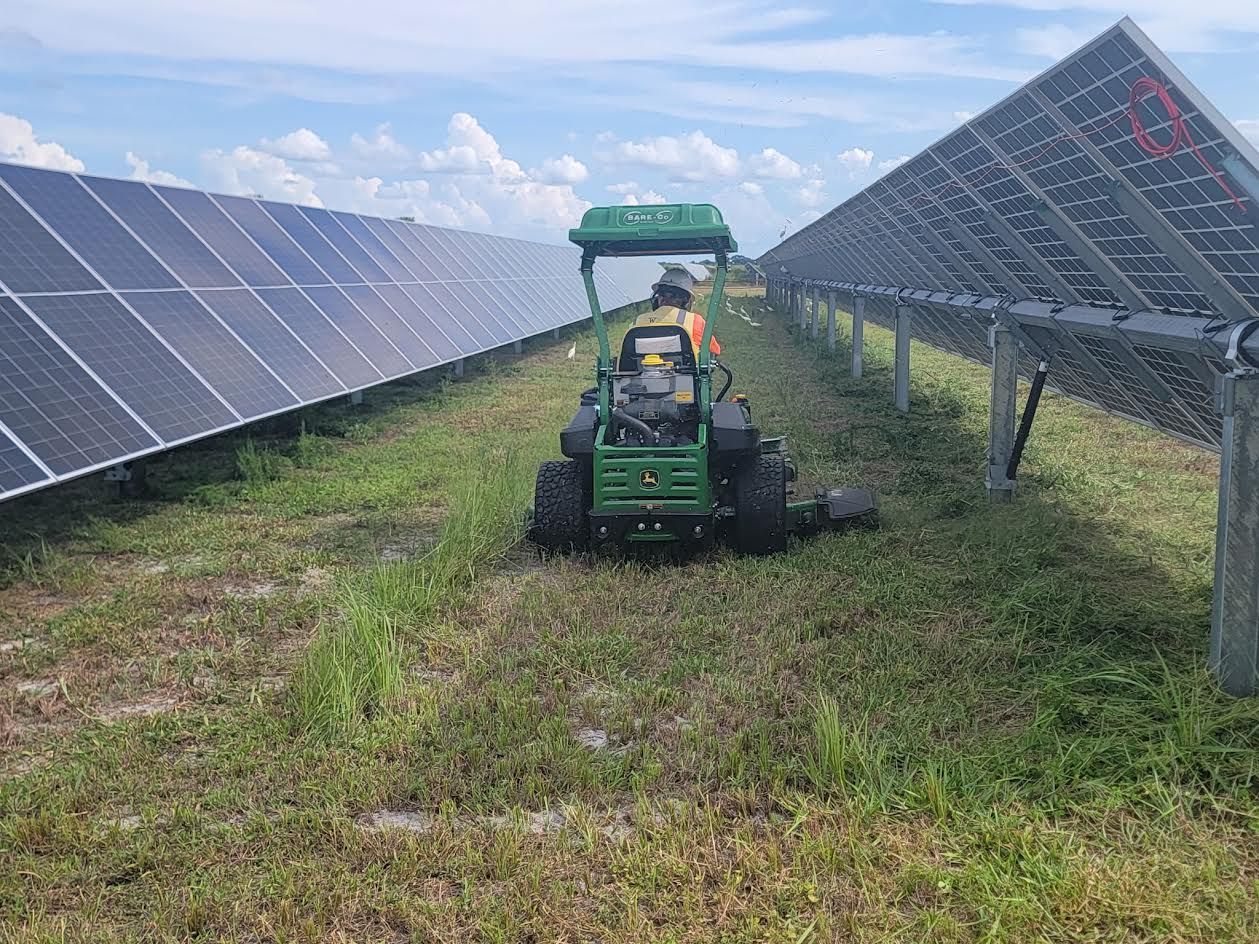Green tractor mowing grass between rows of solar panels on a sunny day