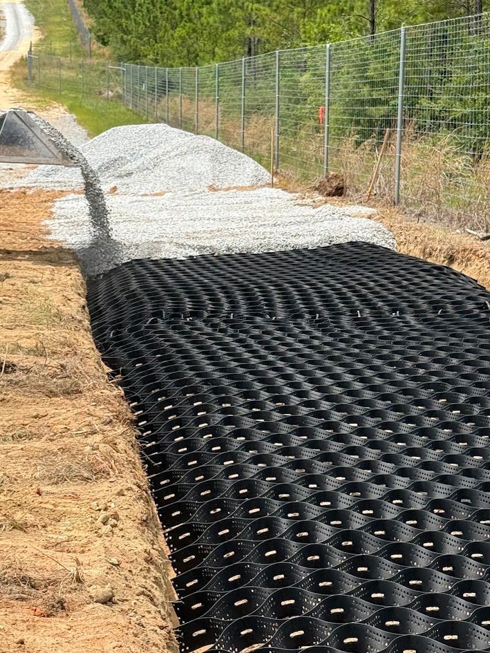 Black geocell mat laid on a gravel path beside a chain-link fence in a wooded area