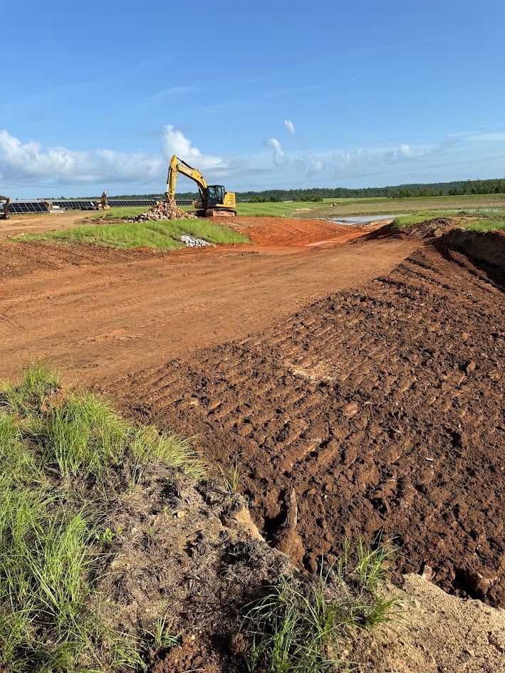 Excavator grading a dirt field under a blue sky, with piles of soil and a rural landscape.