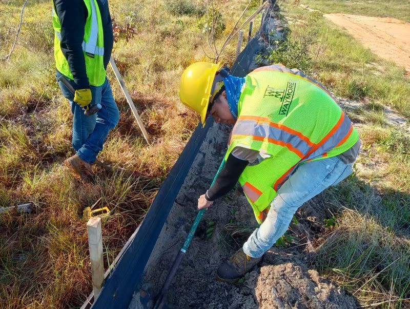 Two workers in safety vests and hard hats inspect a black pipe beside a dirt path.