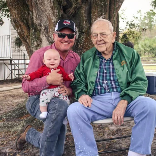 Three people sit on a tree swing outdoors, smiling under a large tree.