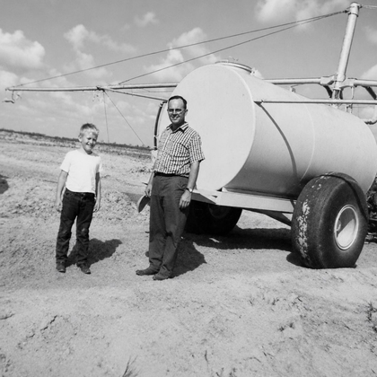 Two men standing beside a large cylindrical tank trailer on a dirt road in a rural field