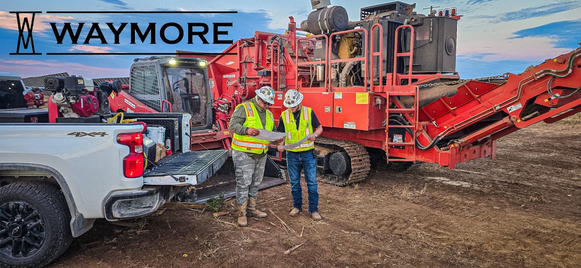 Two workers review a map in a logging site with a yellow wood chipper and stacked debris.