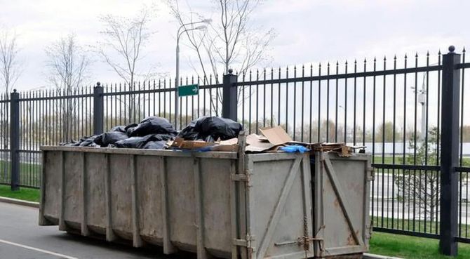 Large dumpster full of trash beside a black metal fence on a cloudy day