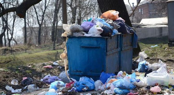 Blue dumpster overflowing with trash bags and litter in a bare outdoor lot