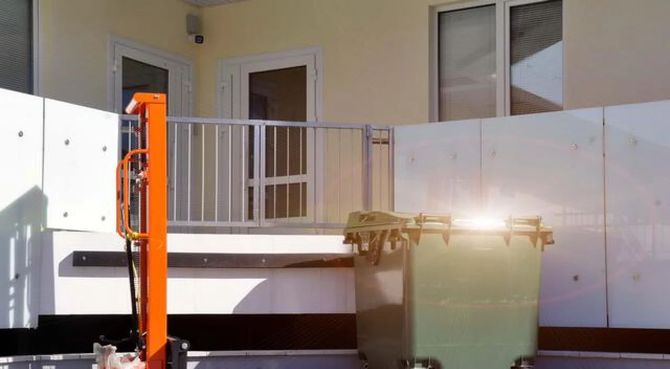 Outdoor patio with white cabinets, an orange lift, and a green dumpster in front of a building