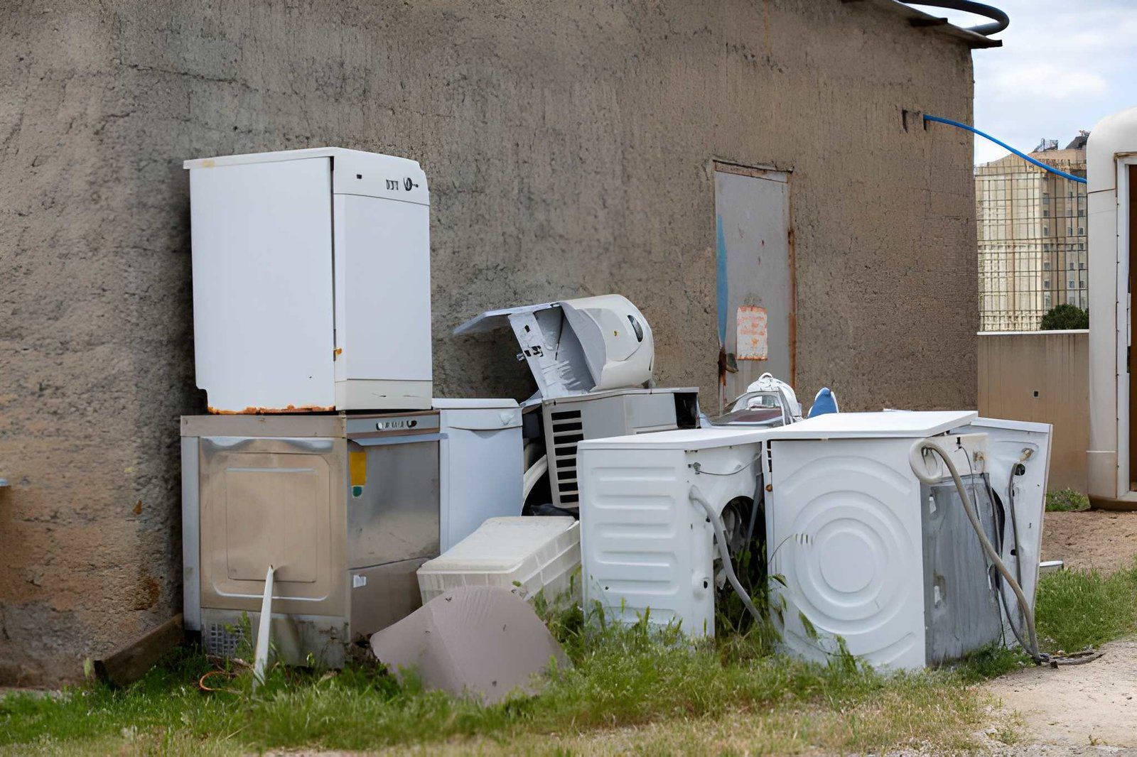 Assorted old appliances and metal scraps piled beside a weathered wall in a grassy yard