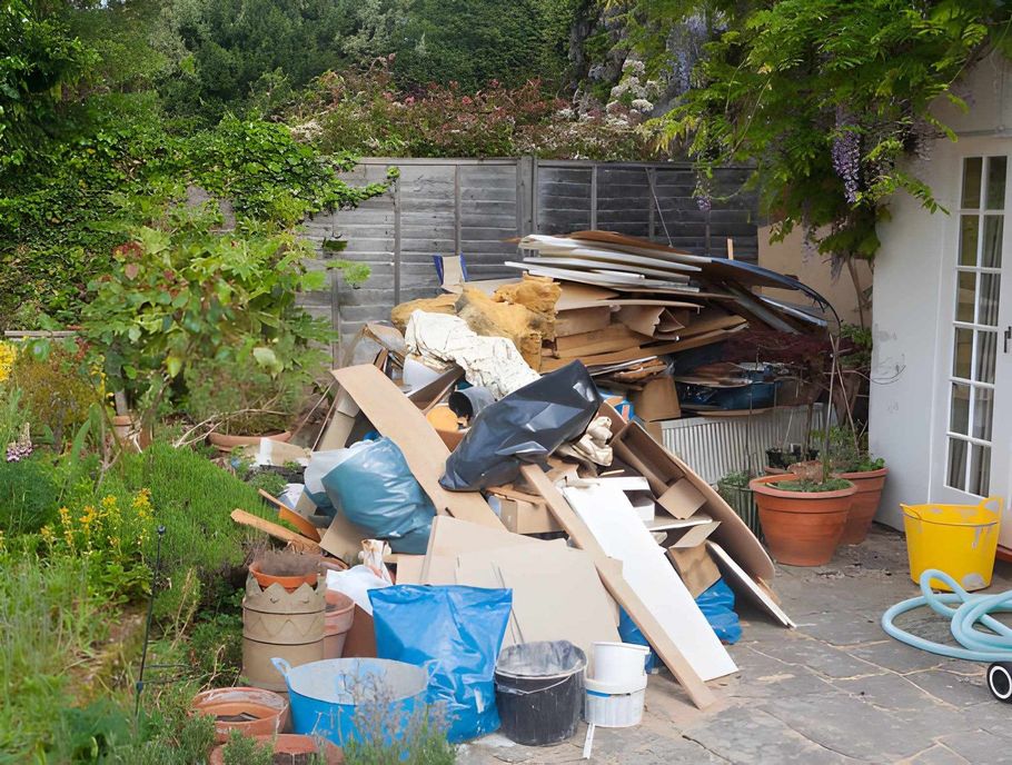 Outdoor patio cluttered with stacked boards, bins, and garden tools beside a house wall