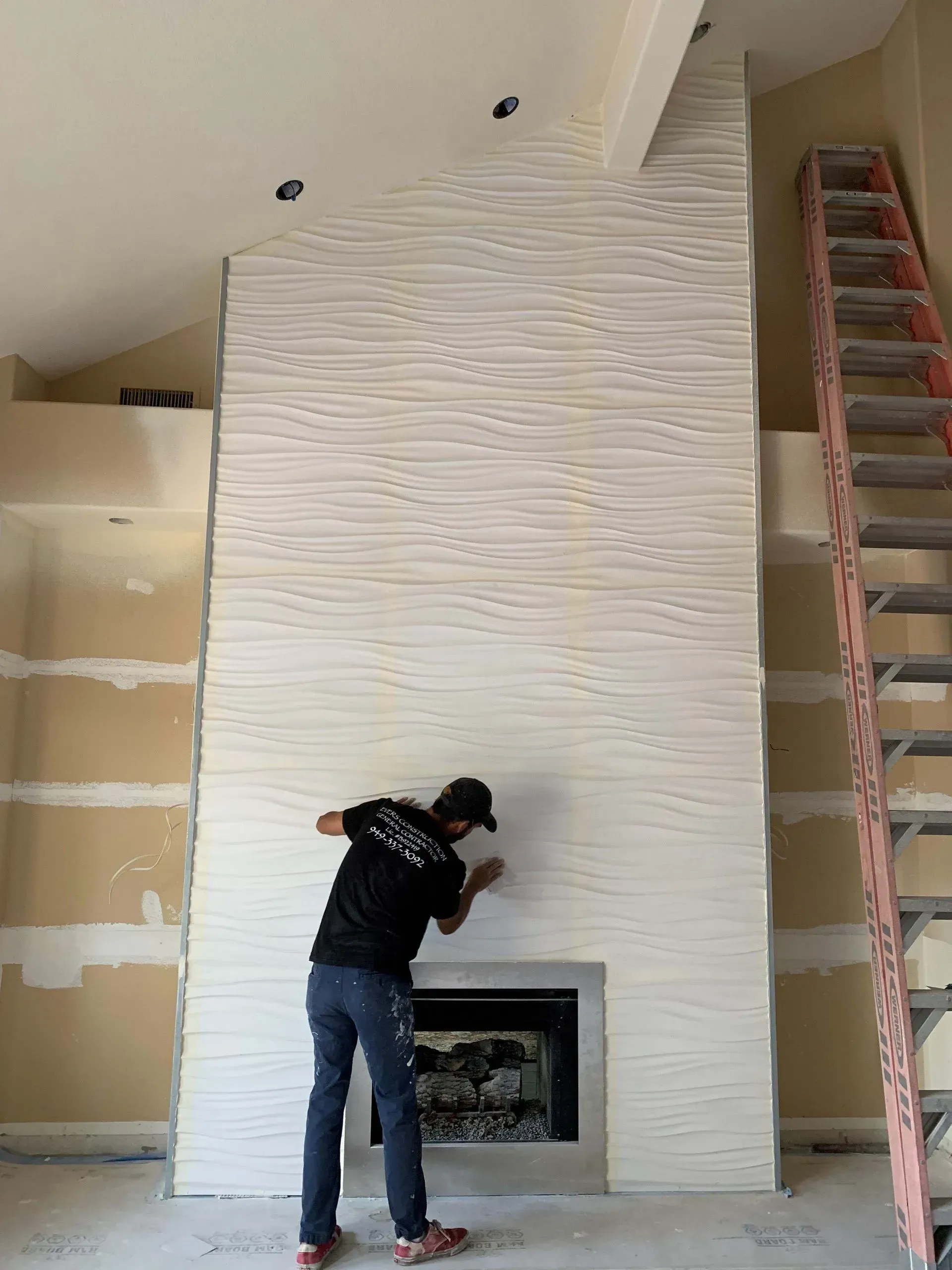 Man working on white textured fireplace in unfinished room, ladder to the side.