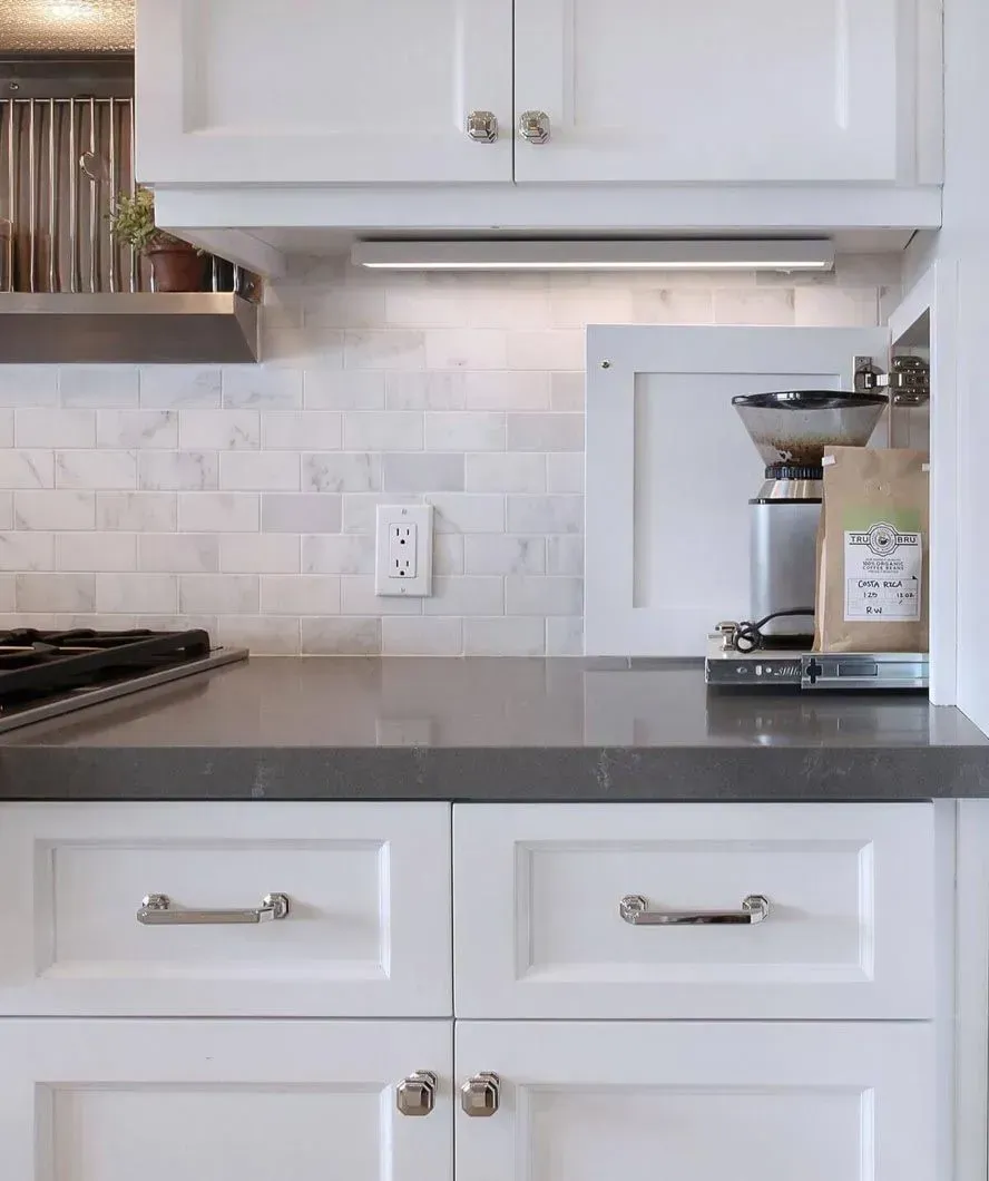 White kitchen cabinets with gray countertop, tile backsplash, and overhead lighting.