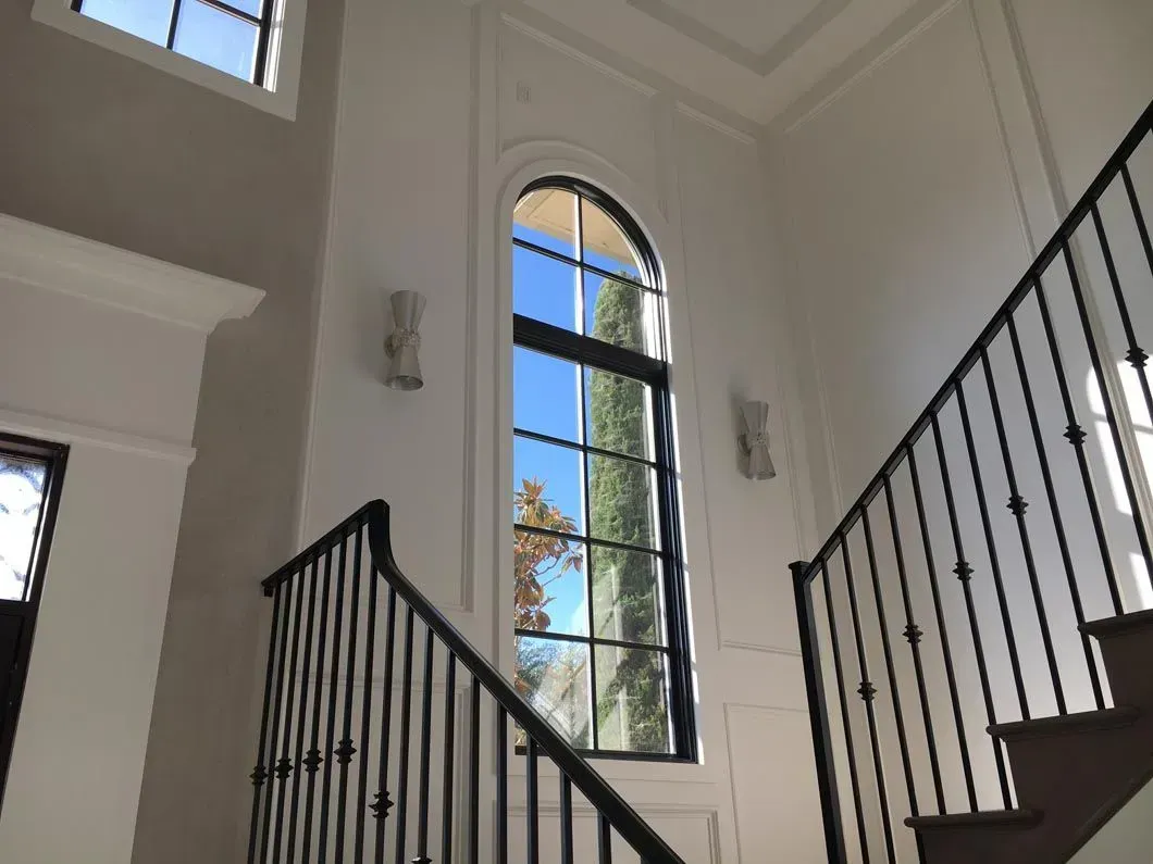 Two-story foyer with staircase and arched window, black railing, white walls, and bright sky reflected in the window.