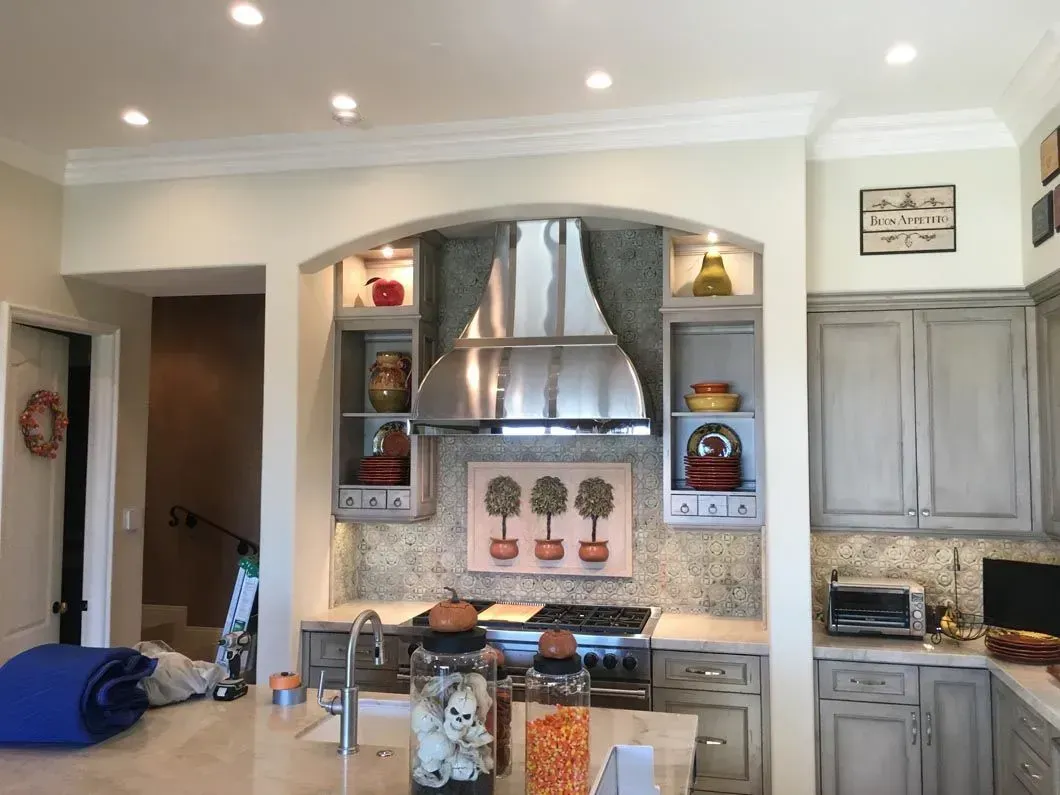 Kitchen with stainless steel range hood, built-in shelves, and gray cabinets. Countertops and recessed lighting are visible.