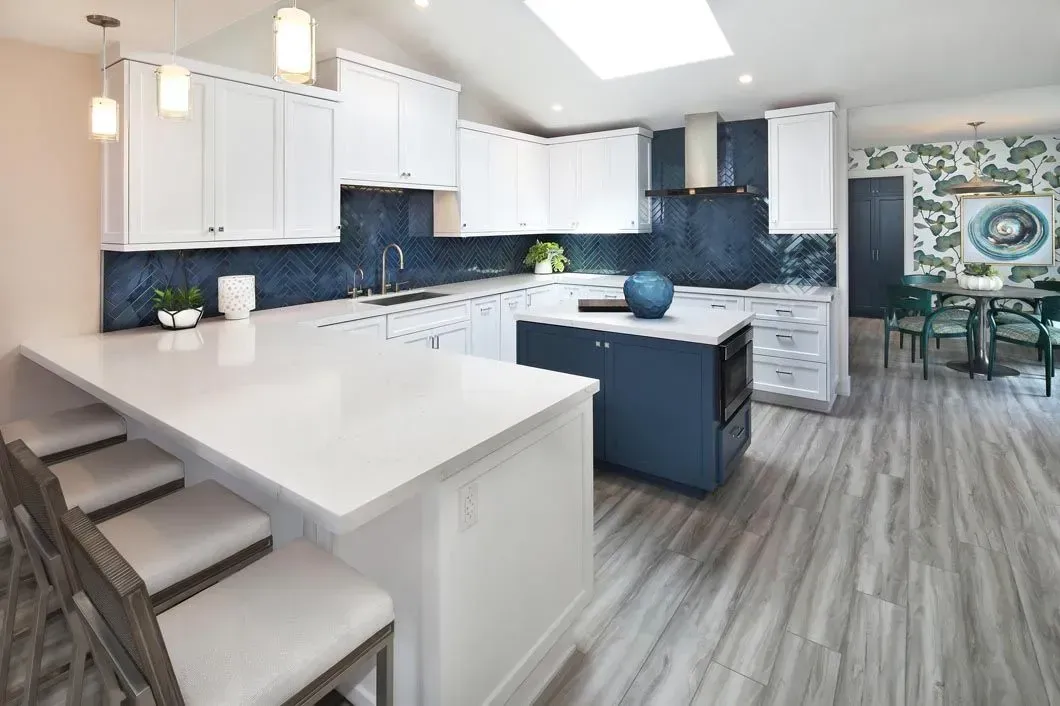 Modern kitchen with white cabinets, blue backsplash, island, and dining area with floral wallpaper.