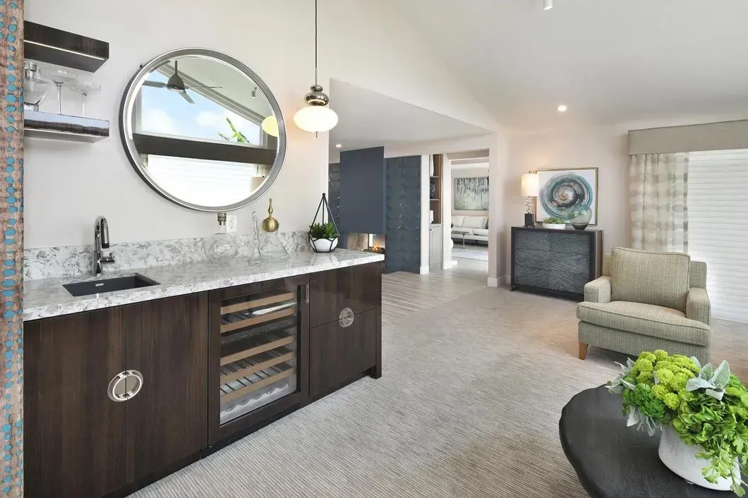A bar area with a wine fridge, sink, and round mirror. A light-colored carpet and armchair sit in the lounge.