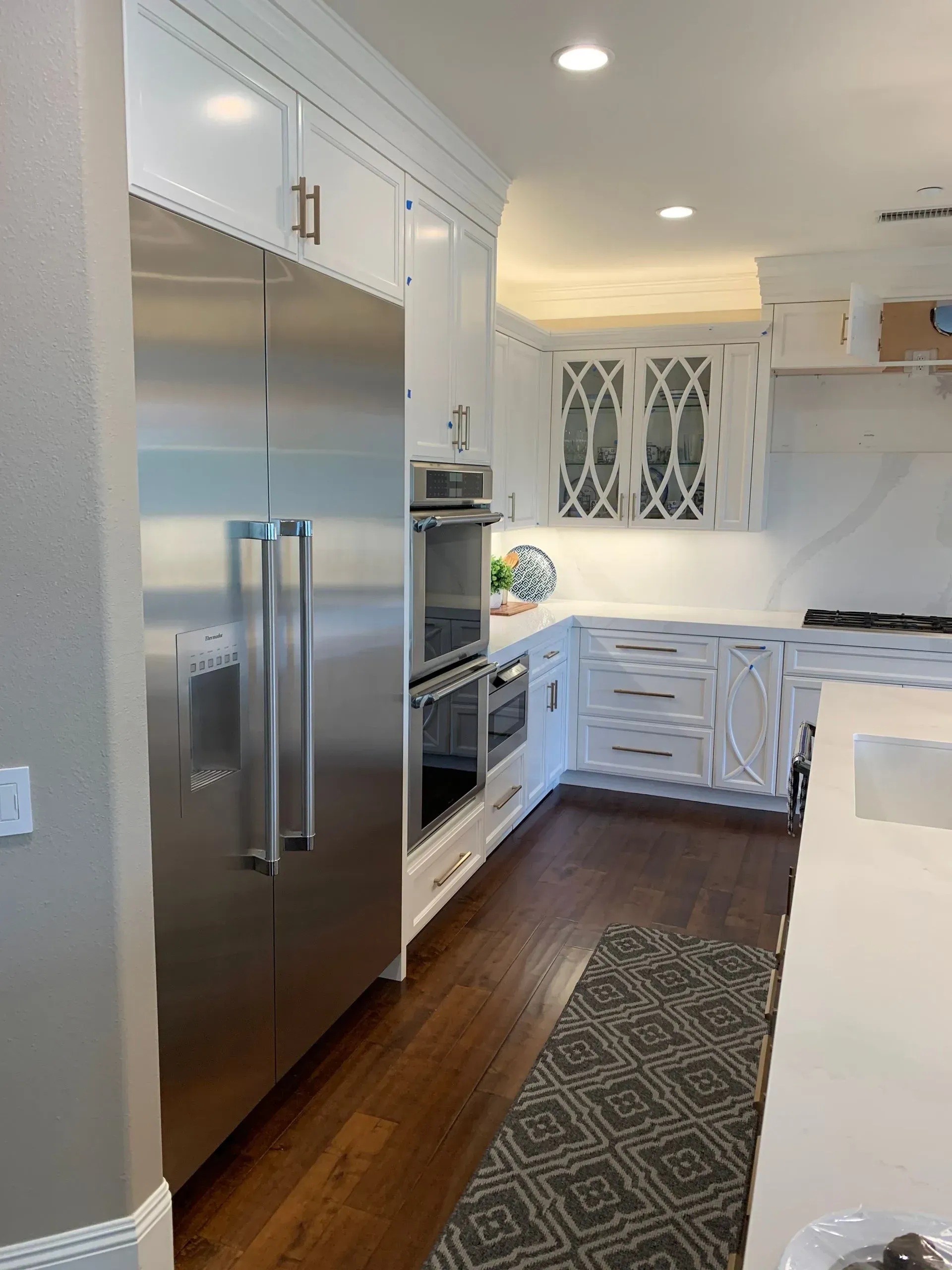 Stainless steel refrigerator and oven in a bright white kitchen with wood floors and a patterned rug.