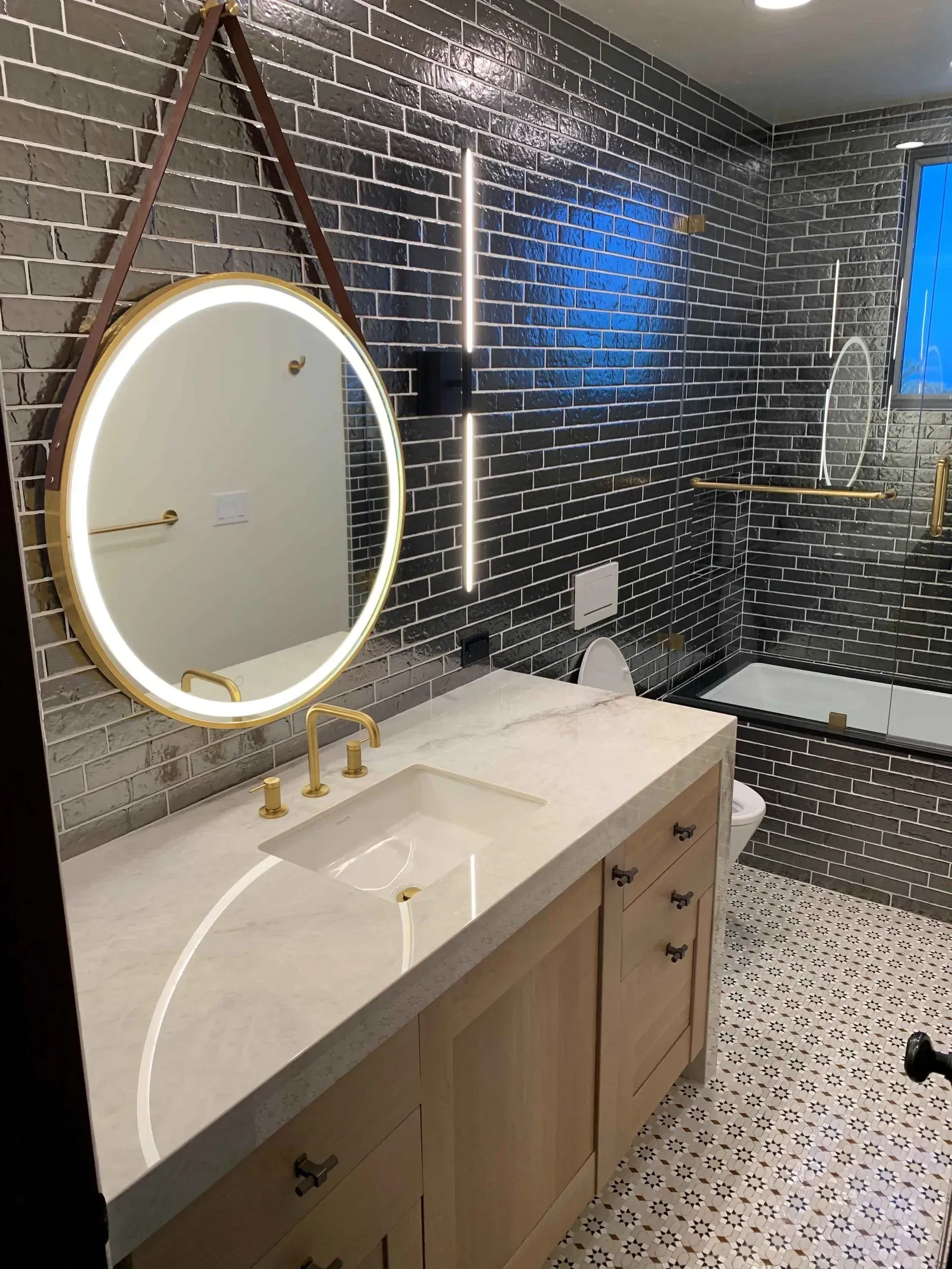 Bathroom with gold fixtures, white countertop, and gray tiled walls and patterned floor; mirror with light.