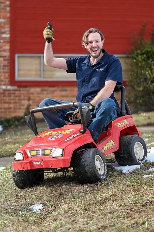 A man is sitting on a toy lawn mower in front of a red building.