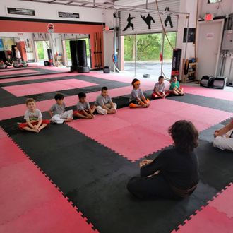 Lil Kickers seated in a martial arts studio, listening to an instructor on red and black mats.
