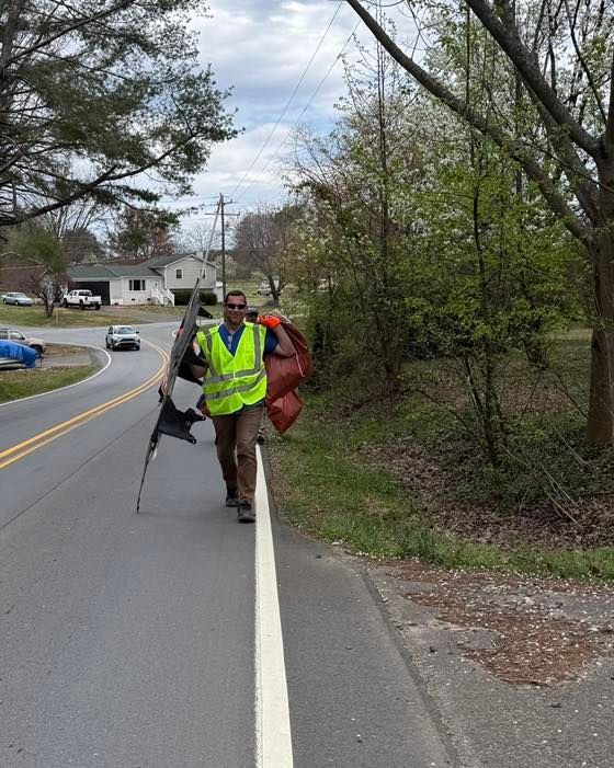 Two people walking along a roadside, one in a neon safety vest carrying a tripod.