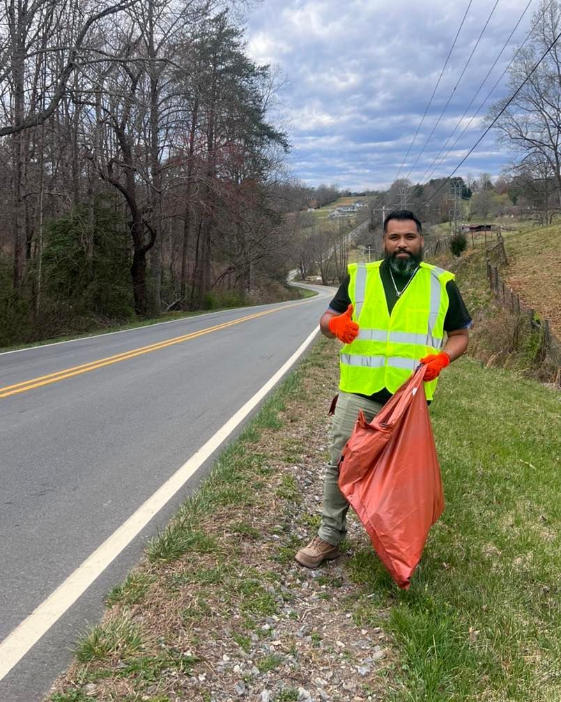 Roadside cleanup worker in neon vest and orange gloves holding a trash bag beside a rural road.
